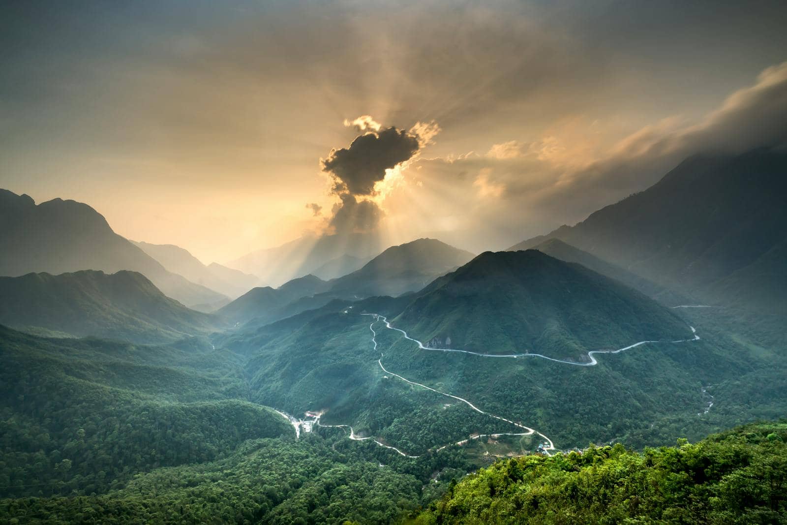 Sun rays break through clouds over a winding road through green mountains